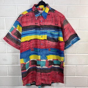 Può includere: Camicia a maniche corte con un colorato design astratto. La camicia presenta un mix di strisce orizzontali e motivi verticali rossi, blu, gialli e marroni. Il colletto e la tasca sono visibili. La camicia è appesa a una gruccia nera.