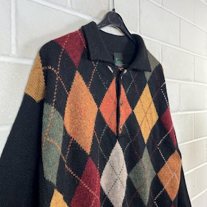 K&ouml;nnte beinhalten: Ein schwarzer Argyle-Pullover mit Kragen und Knopfleiste. Der Pullover hat ein klassisches Rautenmuster in Rot-, Orange-, Gelb-, Gr&uuml;n- und Graut&ouml;nen. Die &Auml;rmel und der Kragen sind einfarbig schwarz.