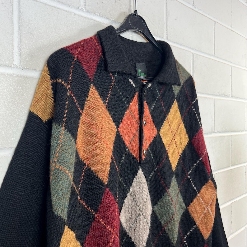 K&ouml;nnte beinhalten: Ein schwarzer Argyle-Pullover mit Kragen und Knopfleiste. Der Pullover hat ein klassisches Rautenmuster in Rot-, Orange-, Gelb-, Gr&uuml;n- und Graut&ouml;nen. Die &Auml;rmel und der Kragen sind einfarbig schwarz.