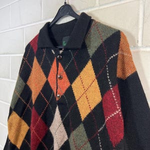 K&ouml;nnte beinhalten: Ein schwarzer Argyle-Pullover mit Kragen und drei braunen Kn&ouml;pfen. Der Pullover hat ein klassisches Rautenmuster in Orange-, Rot-, Gr&uuml;n- und Beiget&ouml;nen. Der Pullover h&auml;ngt an einem schwarzen Kleiderb&uuml;gel vor einer wei&szlig;en Backsteinmauer.