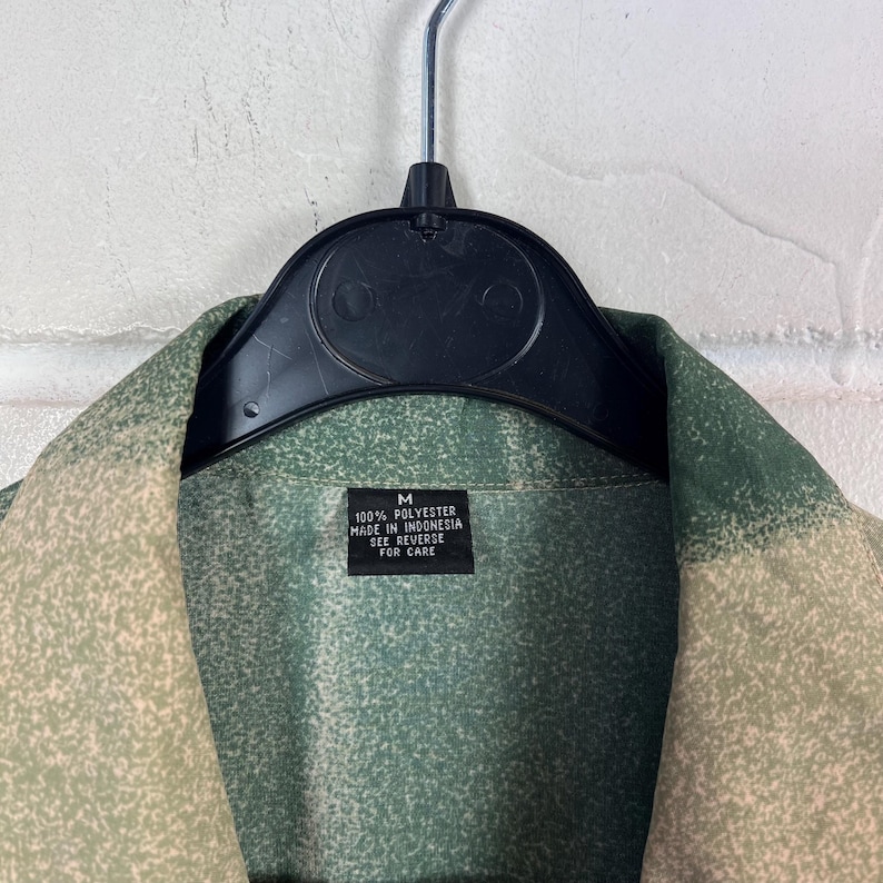 K&ouml;nnte beinhalten: Ein gr&uuml;n-beiges Hemd mit Kn&ouml;pfen, das an einem schwarzen Kleiderb&uuml;gel h&auml;ngt. Das Hemd hat ein vertikales Streifenmuster. Ein Etikett auf dem Hemd lautet "M 100% Polyester Made in Indonesia See Reverse for Care."