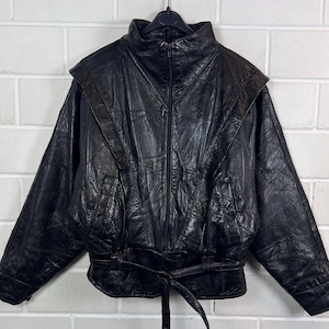 Könnte beinhalten: Eine schwarze Lederjacke mit hohem Kragen, durchgehendem Frontreißverschluss und langen Ärmeln. Die Jacke ist aus mehreren Paneelen aus strukturiertem Leder gefertigt und verfügt über einen passenden Ledergürtel in der Taille.