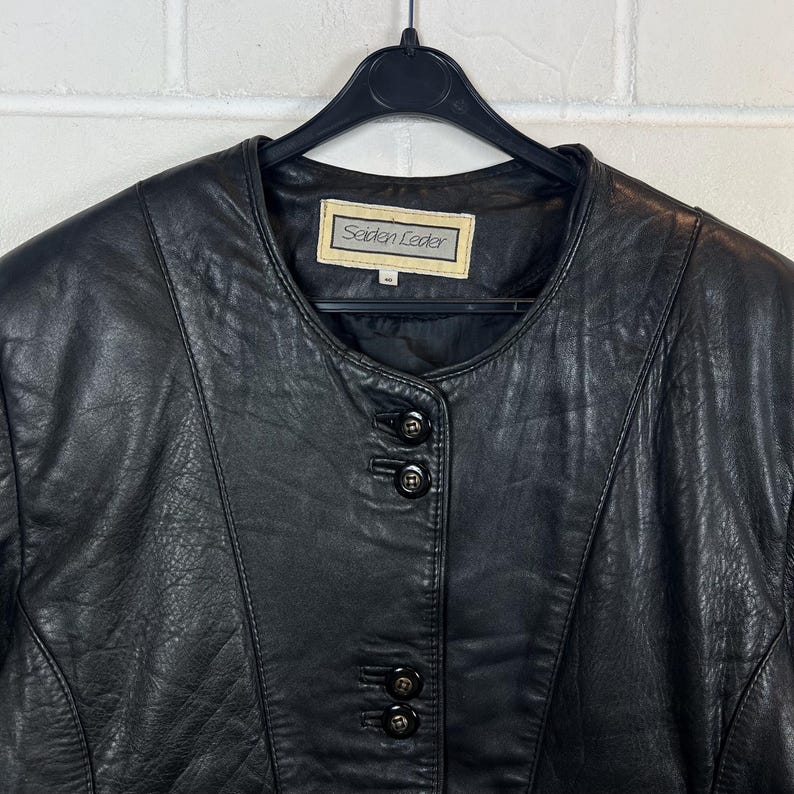 Op de afbeelding: Zwarte leren jas met een ronde kraag en een sluiting met drie knopen. De jas hangt aan een zwarte hanger. Het label luidt "Seiden Leder".