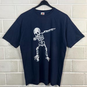 Könnte beinhalten: Marineblaues T-Shirt mit einem weißen Skelett, das dabbt und einen Fußball hält. Das T-Shirt hat kurze Ärmel und ist aus einem weichen Material. Das T-Shirt hängt an einem schwarzen Kleiderbügel.