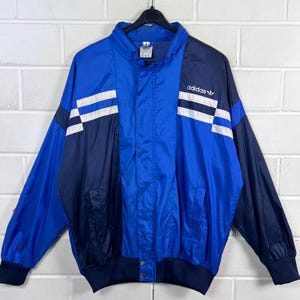 May include: A vintage Adidas track jacket in blue and navy, featuring white stripes on the sleeves. The jacket has a full zip, a stand-up collar, and the Adidas logo. It is displayed on a black hanger.