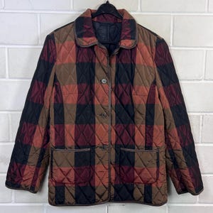 May include: A quilted jacket featuring a large-scale plaid pattern in shades of black, brown, and deep red. The jacket has a classic collar, long sleeves, three front buttons, and two patch pockets at the waist, all finished with dark trim.