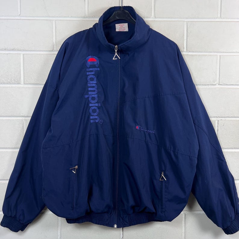 May include: Navy blue Champion jacket with a full zipper, a stand-up collar, and two front pockets. The Champion logo is embroidered in red on the chest and vertically down the left side. The jacket has elastic cuffs and hem.