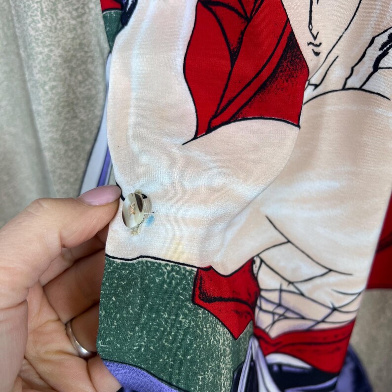 K&ouml;nnte beinhalten: Nahaufnahme eines gemusterten Stoffes mit einem gekn&ouml;pften &Auml;rmel. Der Stoff weist ein Design mit roten, wei&szlig;en und schwarzen Elementen auf. Eine Hand h&auml;lt den &Auml;rmel und zeigt das Knopfd Detail und die Textur des Stoffes.