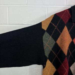 K&ouml;nnte beinhalten: Ein schwarzer Pullover mit einem rautenf&ouml;rmigen Argyle-Muster in Braun-, Orange-, Gr&uuml;n- und Rott&ouml;nen. Der &Auml;rmel ist einfarbig schwarz. Der Pullover hat einen Kragen.