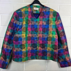 Könnte beinhalten: Eine farbenfrohe, gesteppte Jacke mit einem lebendigen Patchwork-Design. Die Jacke hat einen V-Ausschnitt, einen Knopfverschluss und eine Mischung aus floralen und geometrischen Mustern in Rosa, Blau, Grün und Gelb. Die Jacke hängt an einem schwarzen Kleiderbügel.