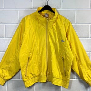 May include: A vintage yellow Adidas jacket with a zip fastening and a small Adidas logo on the left chest. The jacket has a ribbed waistband and cuffs.