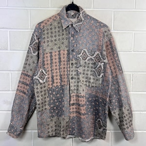 Könnte beinhalten: Ein langärmeliges Hemd mit Patchwork-Design. Das Hemd hat eine Mischung aus Mustern in Grau-, Rosa- und Marineblautönen. Es hängt an einem schwarzen Kleiderbügel vor einer weißen Backsteinmauer.
