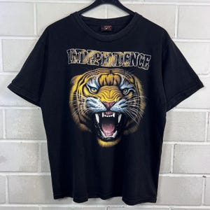 Könnte beinhalten: Ein schwarzes T-Shirt mit kurzen Ärmeln und einer Grafik eines fauchenden Tigerkopfes in Gelb-, Orange- und Weißtönen. Über dem Tiger ist das Wort INDEPENDENCE in einer fetten, weiß umrandeten Schrift gedruckt. Das Shirt hat einen Rundhalsausschnitt.