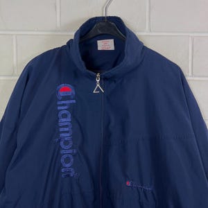 May include: Navy blue Champion jacket with a full zipper and a high collar. The Champion logo is embroidered in red and blue on the left chest. The jacket is hanging on a black hanger against a white brick wall.