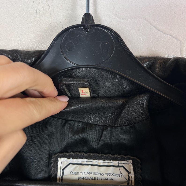 May include: Black leather jacket with a label that reads "L". The jacket is hanging on a black hanger. The label inside the jacket reads "QUESTI CAPI SONO PRODOTTI PARZIALE IN ITALIA".