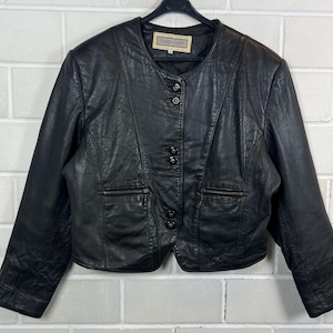 Op de afbeelding: Zwarte leren jas met een ronde kraag, drie knopen en twee zakken met ritssluiting. De jas heeft een label met de tekst "Sophi Leather". De jas hangt aan een zwarte hanger.