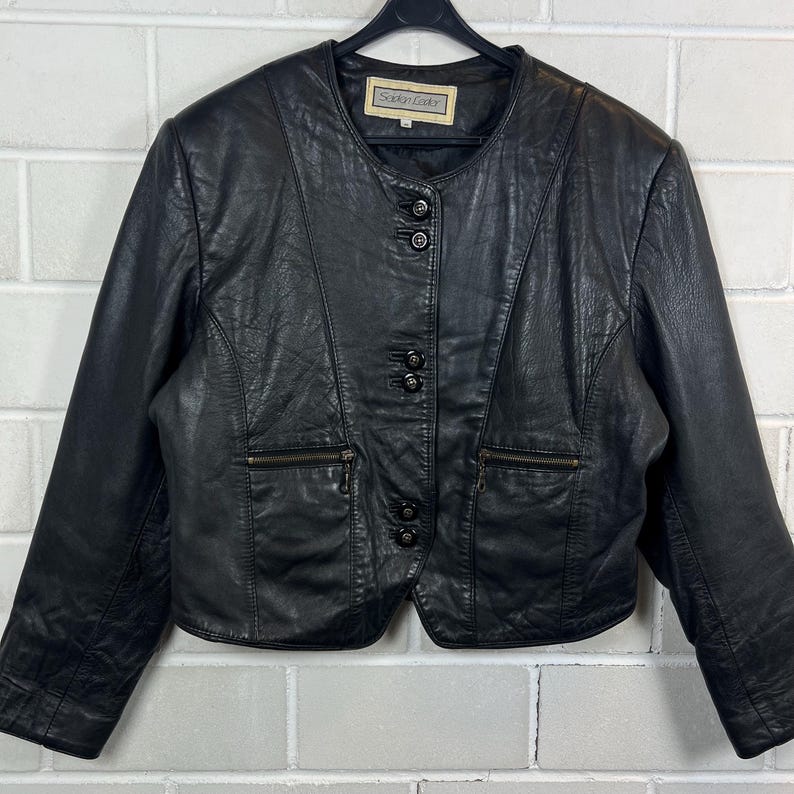 Op de afbeelding: Zwarte leren jas met een ronde kraag, drie knopen en twee zakken met ritssluiting. De jas heeft een label met de tekst "Sophi Leather". De jas hangt aan een zwarte hanger.