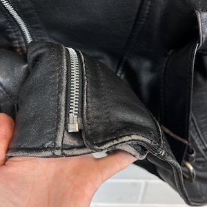 May include: Close-up of a black leather jacket with a silver zipper. The jacket shows signs of wear, with visible creases and a worn appearance around the edges. The zipper is partially open, revealing the interior.