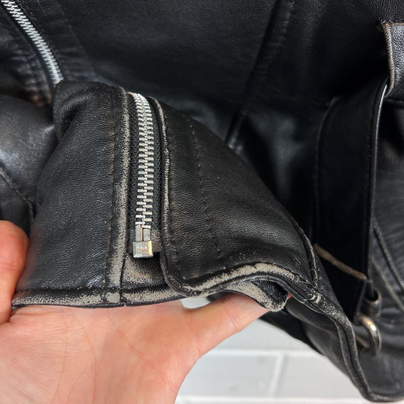 May include: Close-up of a black leather jacket with a silver zipper. The jacket shows signs of wear, with visible creases and a worn appearance around the edges. The zipper is partially open, revealing the interior.