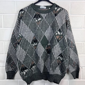 Peut inclure: Un pull vintage vert et gris à motif losanges. Le pull présente un col ras du cou, des poignets et un ourlet côtelés. Les losanges argyle sont dans des tons de vert, de gris et de marron. Le pull est présenté sur un cintre noir contre un mur de briques blanches.