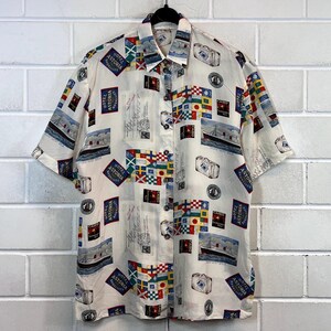 May include: A white short-sleeved button-down shirt with a colourful print of nautical flags, luggage, and the text "Hotel Astoria" repeated throughout.