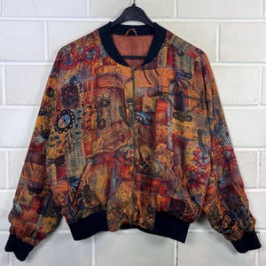 Op de afbeelding: Een bomberjack met lange mouwen en een levendige, veelkleurige patchworkprint in tinten oranje, rood, geel en zwart. Het jack is voorzien van een zwarte geribbelde kraag, boorden en zoom, en heeft een centrale metalen ritssluiting.