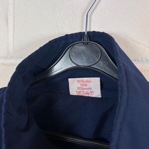 May include: Close-up of a navy blue garment hanging on a black hanger. A small rectangular tag with text is visible. The tag reads "100% Microfibre FUTTER 100% Baumwolle" with care symbols.