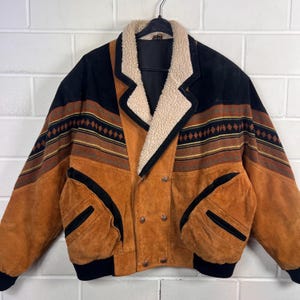 May include: A vintage suede jacket with a shearling collar. The jacket features a colour block design with black, brown, and tan panels. The sleeves have a geometric pattern. The jacket has two front pockets and a double-breasted closure.
