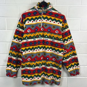 Könnte beinhalten: Ein farbenfroher Fleece-Pullover mit hohem Kragen und einem Viertel-Reißverschluss. Das Muster zeigt geometrische Formen in Rot, Gelb, Blau, Grün und Weiß. Das langärmlige Oberteil hängt an einer weißen Backsteinmauer.