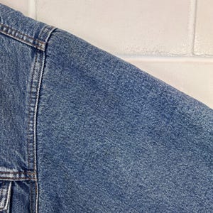 以下が含まれることがあります： 青いデニムジャケットのクローズアップ。画像は肩と袖の一部を示しており、ステッチと縫い目が見えます。デニムはクラシックで質感のある外観をしています。