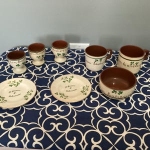 May include: A set of Irish-themed ceramic mugs, egg cups, and a bowl. The mugs and egg cups have green shamrock designs and the text "From Killarney", "From Bahe" and "From Gaelic" on them. The bowl has a green shamrock design and the text "From Killarney" on it. The items are on a blue and white patterned tablecloth.