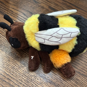 May include: A plush bee toy featuring a brown head with black antennae and round black eyes. The body has alternating horizontal stripes of bright yellow and black plush fabric. It includes two off-white wings with stitched vein details.