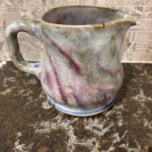 Könnte beinhalten: Handgefertigter Keramikkrug mit Henkel und Ausguss. Der Krug hat ein marmoriertes Design mit Grau-, Lila- und Blautönen. Der Boden hat ein blaues Band. Perfekt zum Servieren von Getränken.