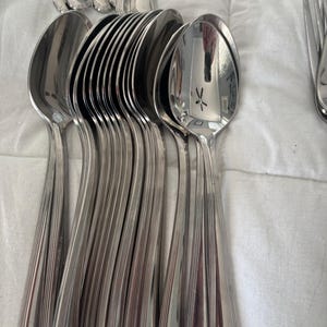 May include: A collection of shiny, silver spoons arranged on a white surface. The spoons have a classic design with a rounded bowl and a handle featuring vertical lines. The spoons are reflective, showing the surrounding environment.