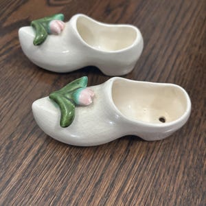 Puede incluir: Dos zuecos holandeses de cerámica blanca con un adorno floral verde y rosa. Los zuecos están vacíos y colocados sobre una superficie de madera oscura. Los zuecos son un objeto decorativo.