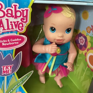 May include: Baby Alive doll in a box. The doll has blonde hair, blue eyes, and is wearing a blue and pink dress. The box reads "Baby Alive" and "Kicks & Cuddles Newborns". The text "TRY ME!" is also visible.