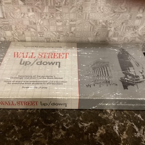 May include: A vintage board game called "Wall Street Up/Down" with a gray and white box featuring a black and white illustration of the New York Stock Exchange building. The game is described as "A game for enterprising minds".