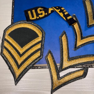 May include: A collection of embroidered military-style patches featuring gold thread on a black background. The set includes a shield-shaped patch with two chevrons, several individual chevron patches, and a black fabric strip with the text U.S. ARMY.