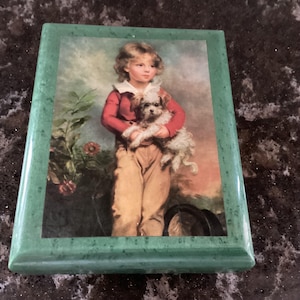 Puede incluir: Una caja de madera verde con una tapa con bisagras. La tapa presenta una pintura vintage de un niño pequeño sosteniendo un perro blanco pequeño. El niño lleva una camisa roja y pantalones marrones.