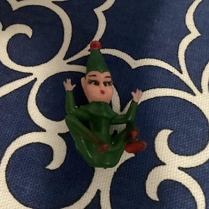 Puede incluir: Una figurita de elfo de plástico verde con un gorro rojo y una cara blanca. El elfo está sentado con las piernas cruzadas y los brazos levantados.