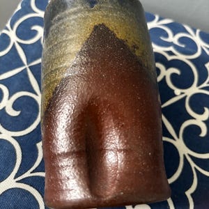 以下が含まれることがあります： ユニークな質感の表面を持つ、手作りのセラミックタンブラー。上部は青と金のブレンドで、下部に向かって濃い茶色に変化しています。円筒形はわずかに先細りになっており、素朴な魅力を添えています。