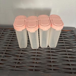 Op de afbeelding: Vier doorzichtige plastic kruidencontainers met lichtroze deksels. De containers zijn rechthoekig met afgeronde bovenkanten en hebben een kleine opening op het deksel. De containers zijn leeg.