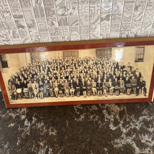 May include: A large, rectangular, black and white panoramic photograph featuring a group of many people posed in front of a building with columns. The photograph is encased in a polished, reddish-brown wooden frame with a thin gold inner border.