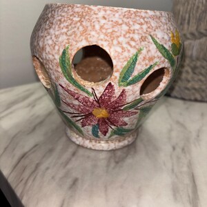 May include: A speckled ceramic vase with a floral design. The vase has a light brown and white speckled exterior, with large holes cut into the sides. The floral design features red and yellow flowers with green leaves. The vase sits on a white surface.