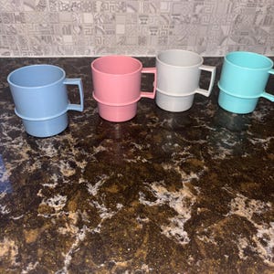 Könnte beinhalten: Vier Kunststoffbecher in Pastellfarben: Hellblau, Rosa, Grau und Türkis. Jeder Becher hat einen Henkel und einen Basisring. Die Becher sind auf einer braunen Arbeitsplatte mit einem gesprenkelten Muster angeordnet, mit einer gemusterten Fliesenspiegel.