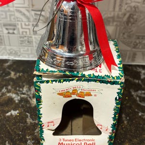 May include: A shiny silver Christmas bell with a red ribbon, resting on a box. The box is white with green and red detailing, and the text "Liberty Bell Christmas" is displayed. The box also reads "3 Tunes Electronic Musical Bell."