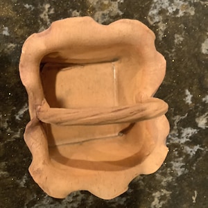 May include: A small, terracotta-colored ceramic basket with a handle. The basket has a scalloped edge and a rough, textured surface.