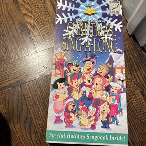May include: A vintage Hanna-Barbera's Christmas Sing-A-Long cassette tape. The cover features cartoon characters singing carols in a snowy scene. The text includes "Special Holiday Songbook Inside!" and "Audio Cassette".