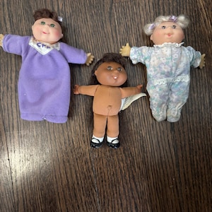 Peut inclure: Trois poupées Cabbage Patch Kids. L'une porte une robe violette, une autre une tenue beige et la troisième un pyjama à fleurs. Toutes les poupées ont des visages en vinyle doux et de petits bras tendus.