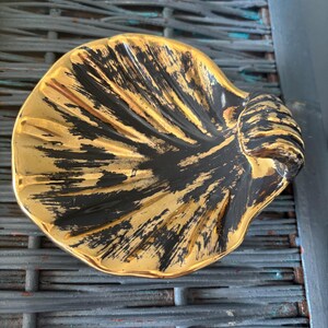 May include: A shell-shaped dish featuring a metallic gold finish with black brushstroke accents radiating from the center. The ceramic piece has a scalloped edge and a textured surface that mimics the natural ridges of a seashell, with a glossy sheen.